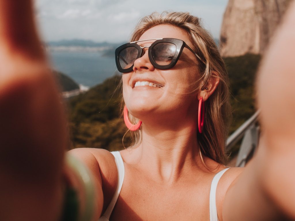 a blonde woman with sunglasses taking a selfie by the ocean while on vacation in order to post it on Instagram