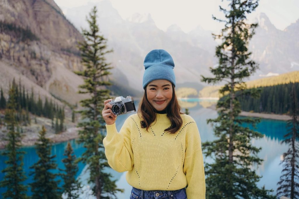 Smiling woman in a light blue beanie and yellow knit sweater standing in front of a lake with her camera, facing the photographer.